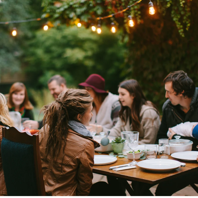 A group of friends gathers around a wooden table for an evening meal outdoors, illuminated by the warm glow of hanging string lights in a garden setting.