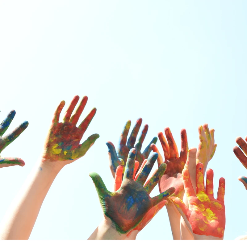 A group of hands covered in splotches of colorful paint are raised up against a clear, light blue sky, suggesting a fun and creative group activity.