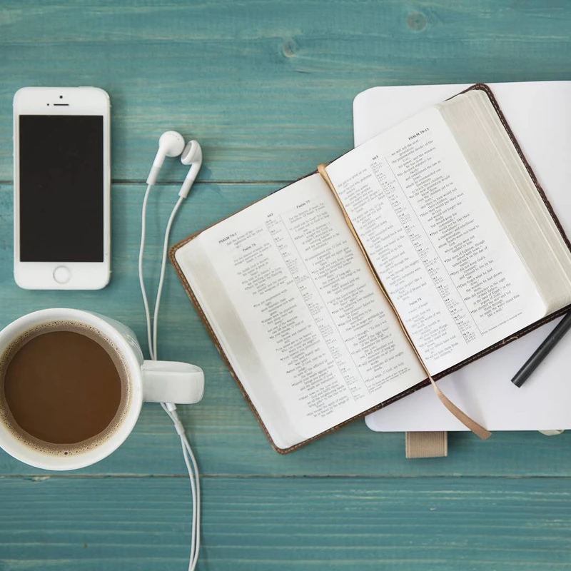 A flat lay image of an open Bible, a smartphone with earbuds, and a mug of coffee on a teal wooden desk, representing a quiet moment of study or worship.