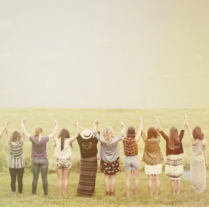 A group of women stand in a line from behind in a grassy field, holding their hands up together in a sign of unity. The photo has a warm, vintage filter.