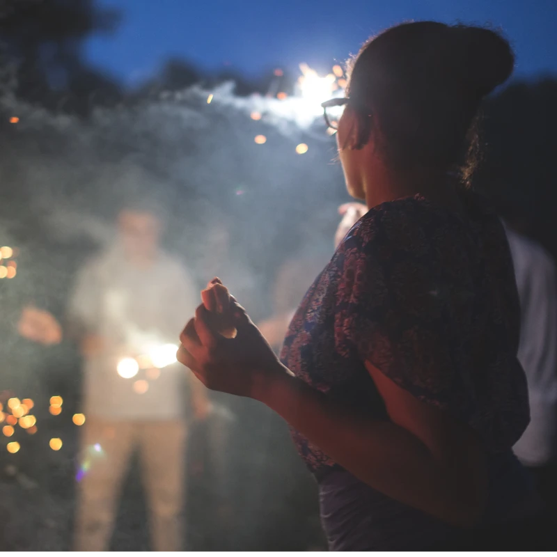 A woman seen from behind watches friends celebrate with lit sparklers at night. The air is filled with smoke and sparks against a dark blue sky.