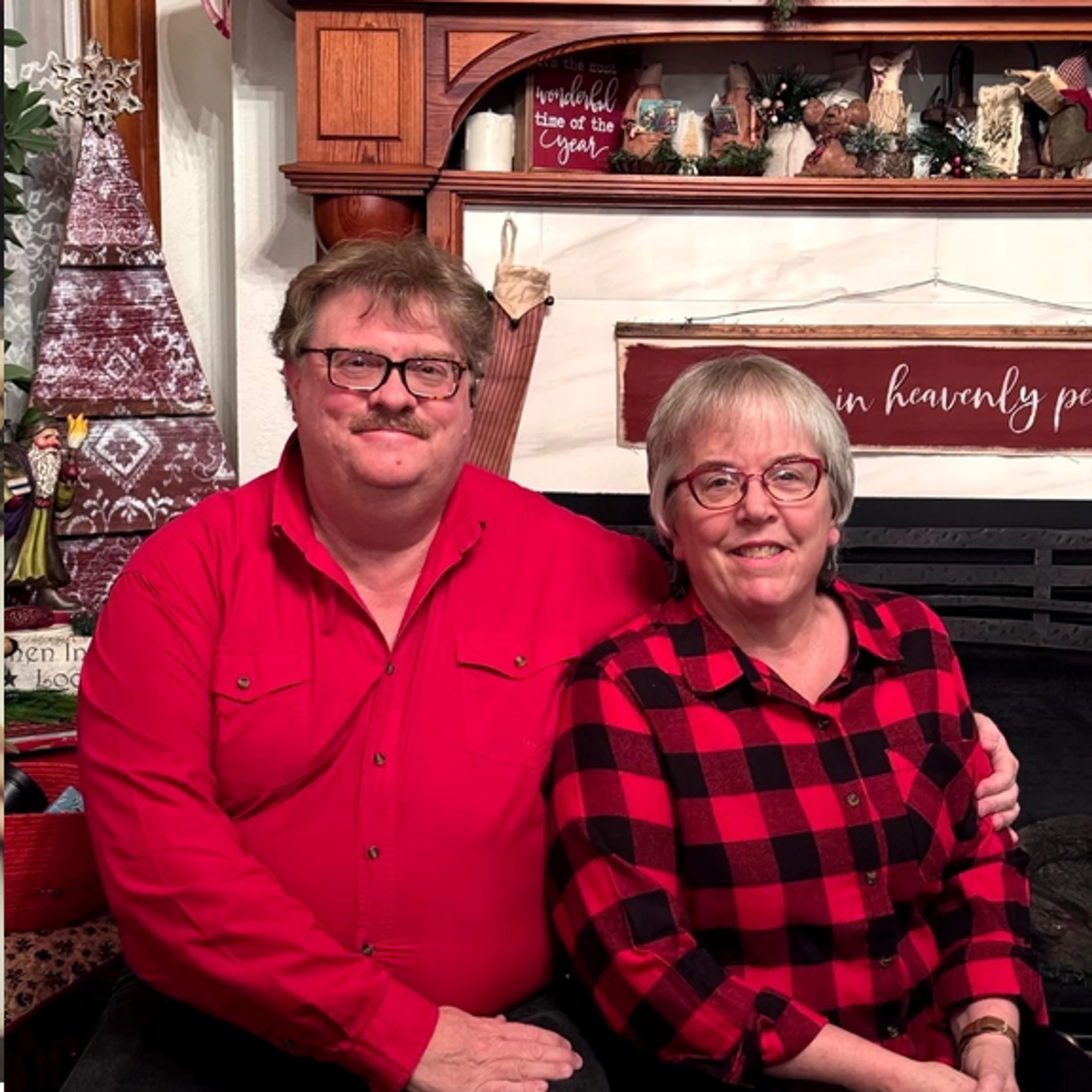 A smiling man in a red shirt and a woman in a red and black plaid shirt sit together in front of a fireplace decorated for the holidays.