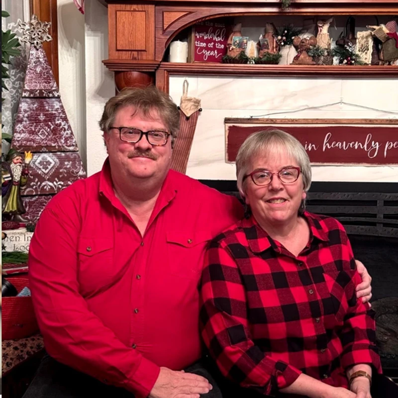 A smiling man in a red shirt and a woman in a red and black plaid shirt sit together in front of a fireplace decorated for the holidays.