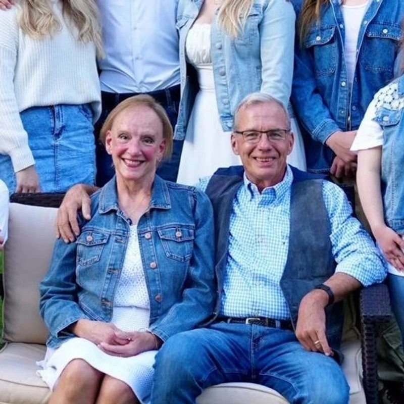 An older couple smiles while sitting on a couch for a group photo. The man has his arm around the woman, and they are both wearing denim and jeans.
