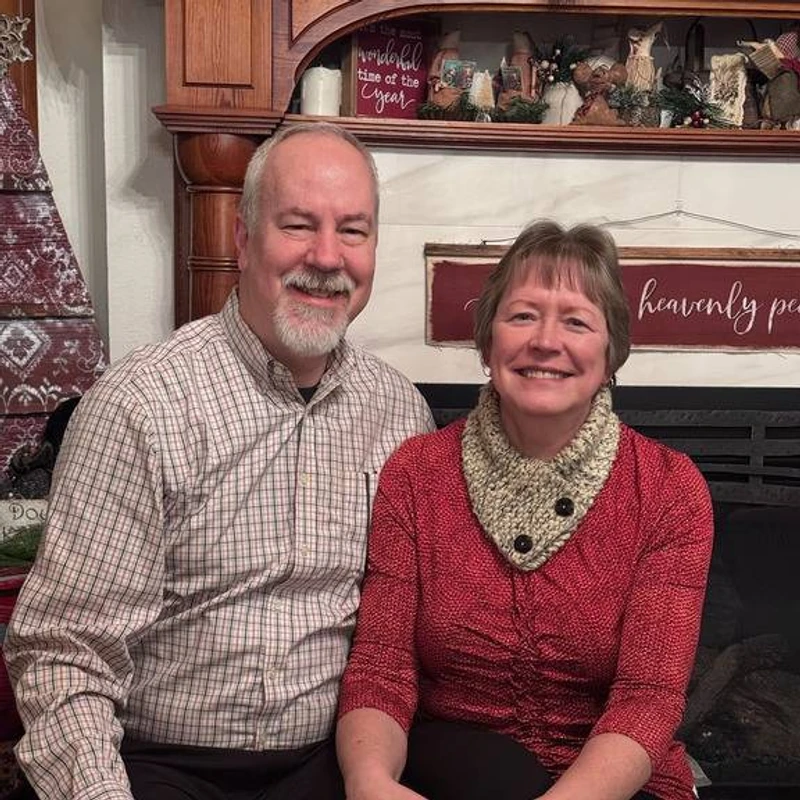 A smiling man and woman sit together in front of a fireplace mantel decorated for the holidays. The man wears a plaid shirt and the woman wears a red top.