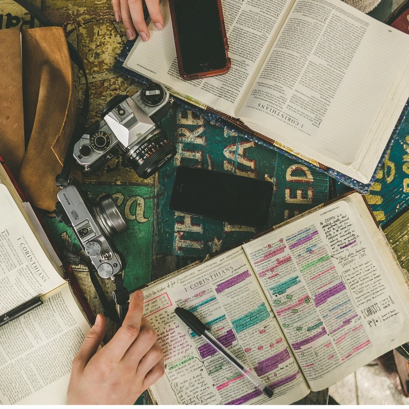 An overhead view of a table with open Bibles, one heavily highlighted and annotated. A hand holds a pen over the page, surrounded by cameras and smartphones.