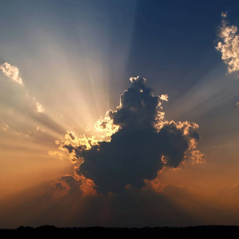 A large, dark cloud obscures the sun, with brilliant golden rays of light fanning out from behind it against a blue and orange sky at sunset.