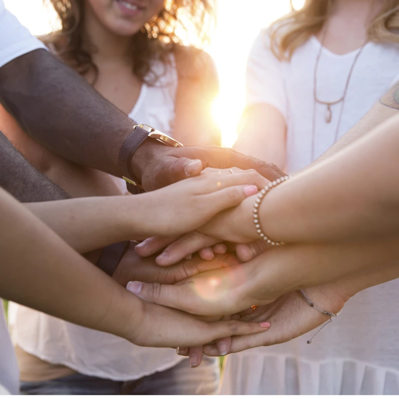 A close-up shot of a diverse group of people stacking their hands together in a symbol of unity and teamwork, illuminated by a warm, setting sun.