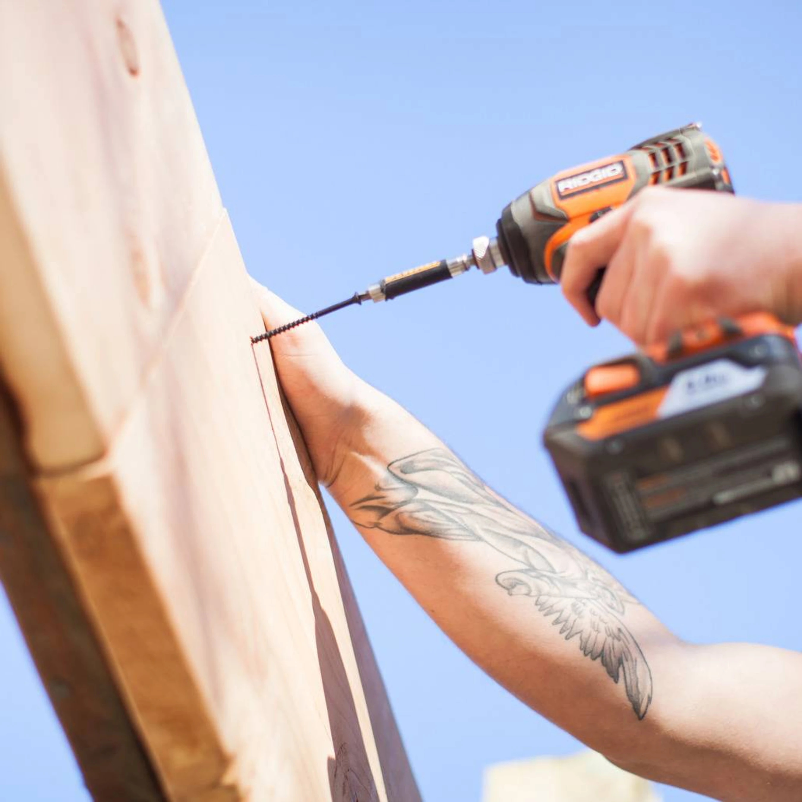 A person with a large tattoo on their arm uses an orange power drill to drive a screw into a wooden beam against a bright blue sky.