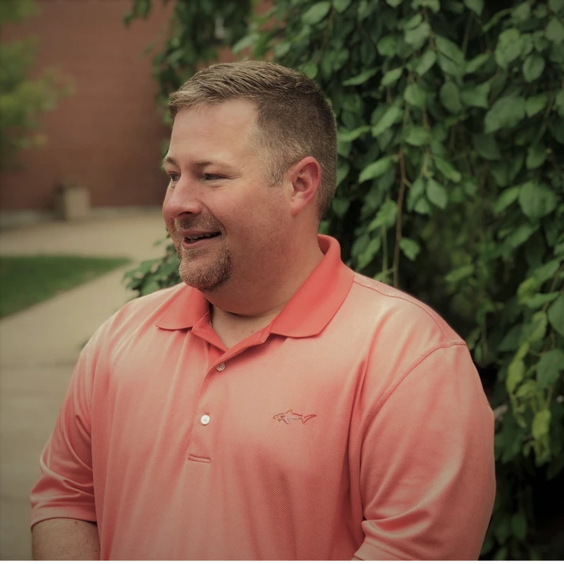 A medium shot of a smiling man with short brown hair and a goatee. He is wearing a coral polo shirt and standing outdoors in front of green leaves.
