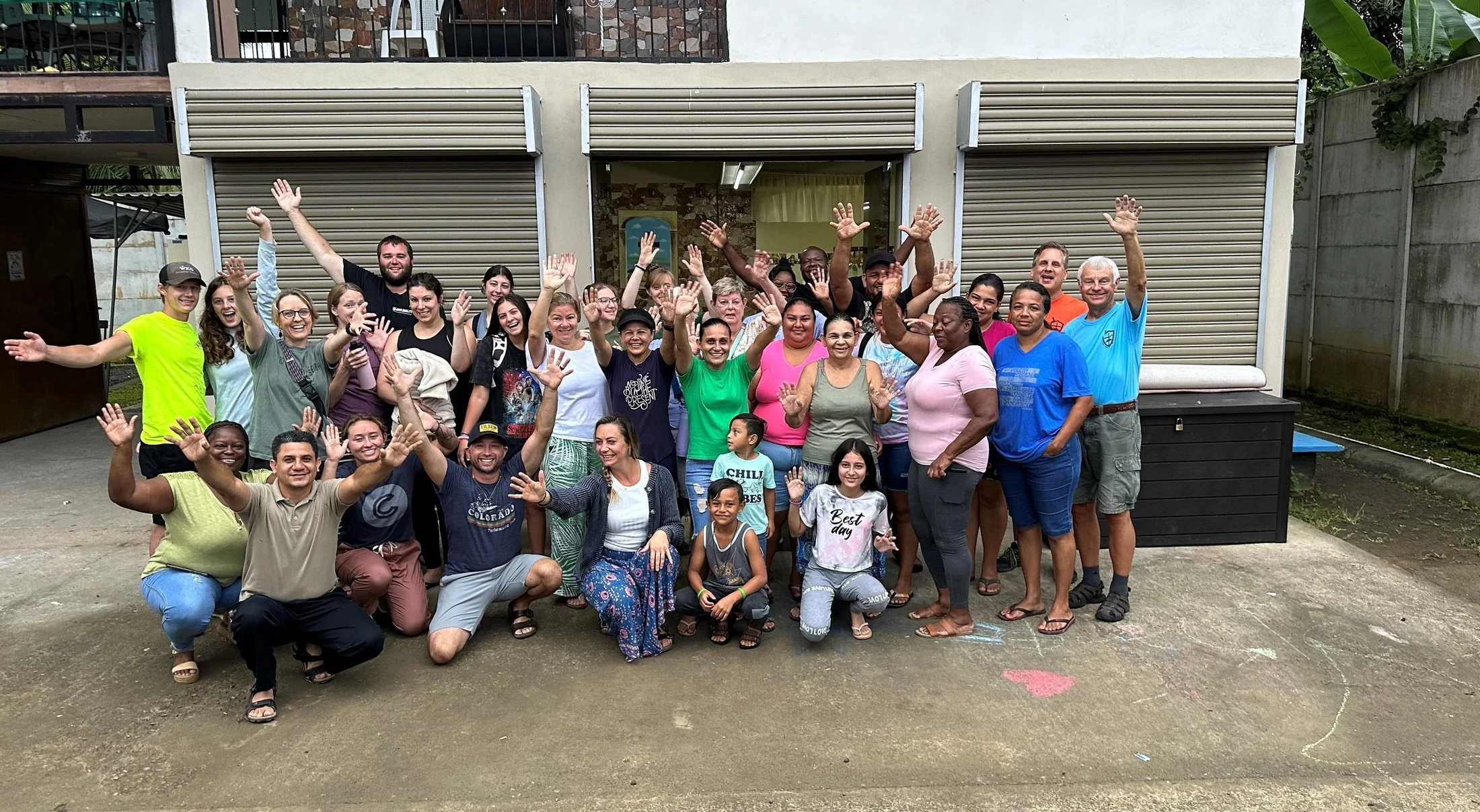 A large, diverse group of adults and children smiling and waving for a group photo outdoors in front of a building.