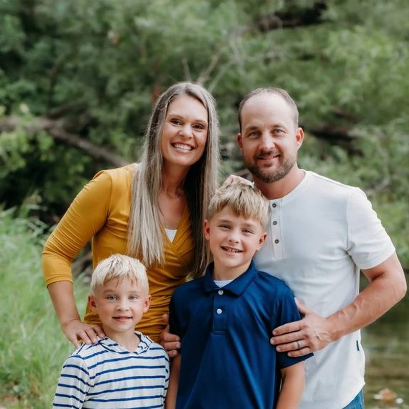 A smiling family of four, a mother and father with their two young sons, posing for a portrait in a park with green trees in the background.
