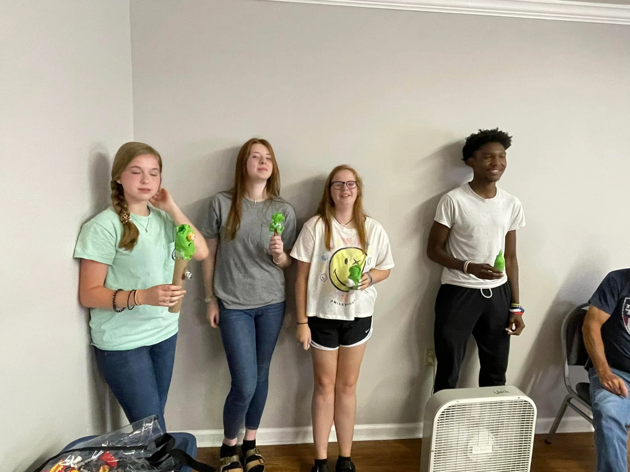 Four teenagers, three girls and one boy, stand in a line against a plain wall inside a room. They are dressed casually and each holds a small green toy.
