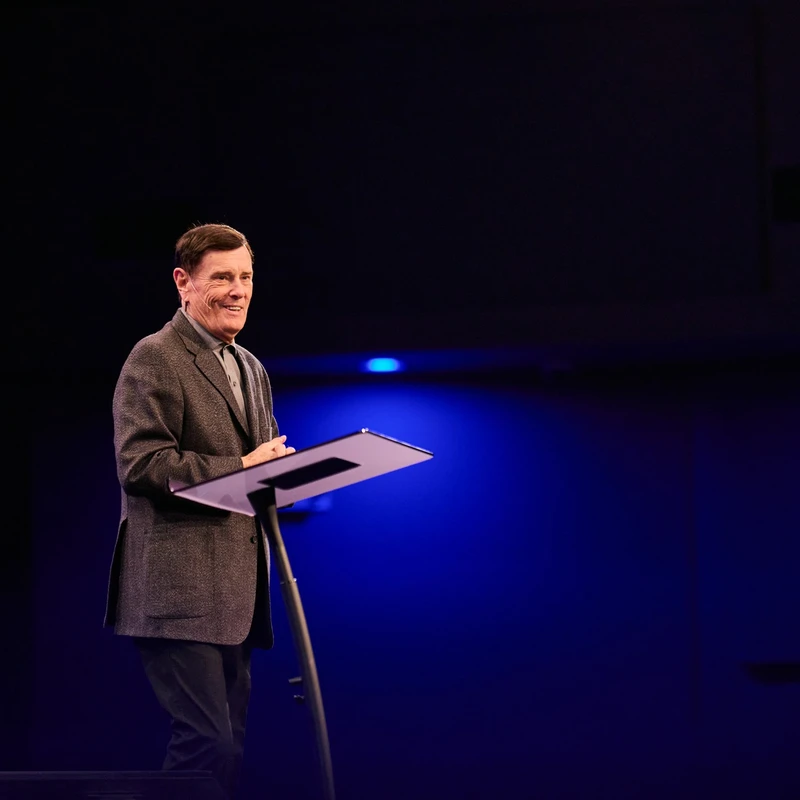 A man with short brown hair, wearing a grey jacket, smiles as he stands at a lectern on a dimly lit stage with blue background lighting.
