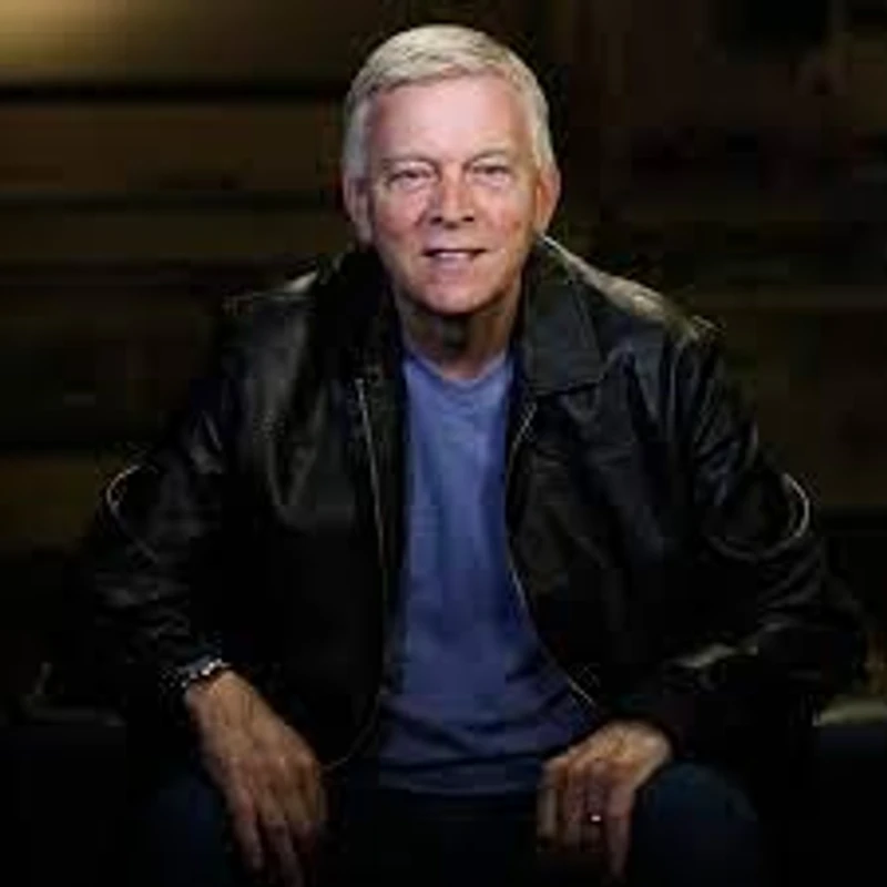A smiling, gray-haired man sits facing the camera. He is wearing a black leather jacket over a blue t-shirt and dark pants in a dimly lit setting.