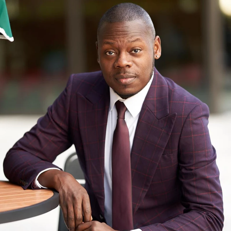 A Black man with a short haircut wearing a purple plaid suit and tie sits at a table outdoors, looking seriously at the camera.