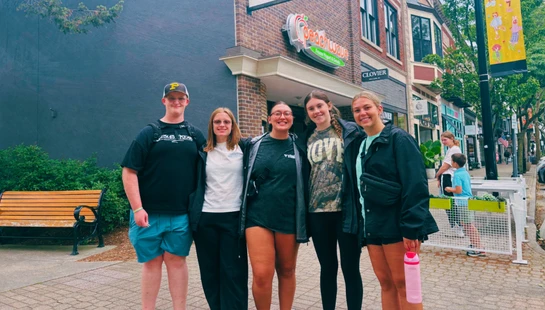 A group of five young adults, one male and four females, stand smiling on a brick sidewalk in front of a Peachwave frozen yogurt shop on a city street.