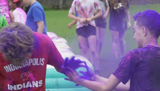Two teenage boys play together outdoors, covered in purple and pink powder during a color festival, with a cloud of purple dust between them.