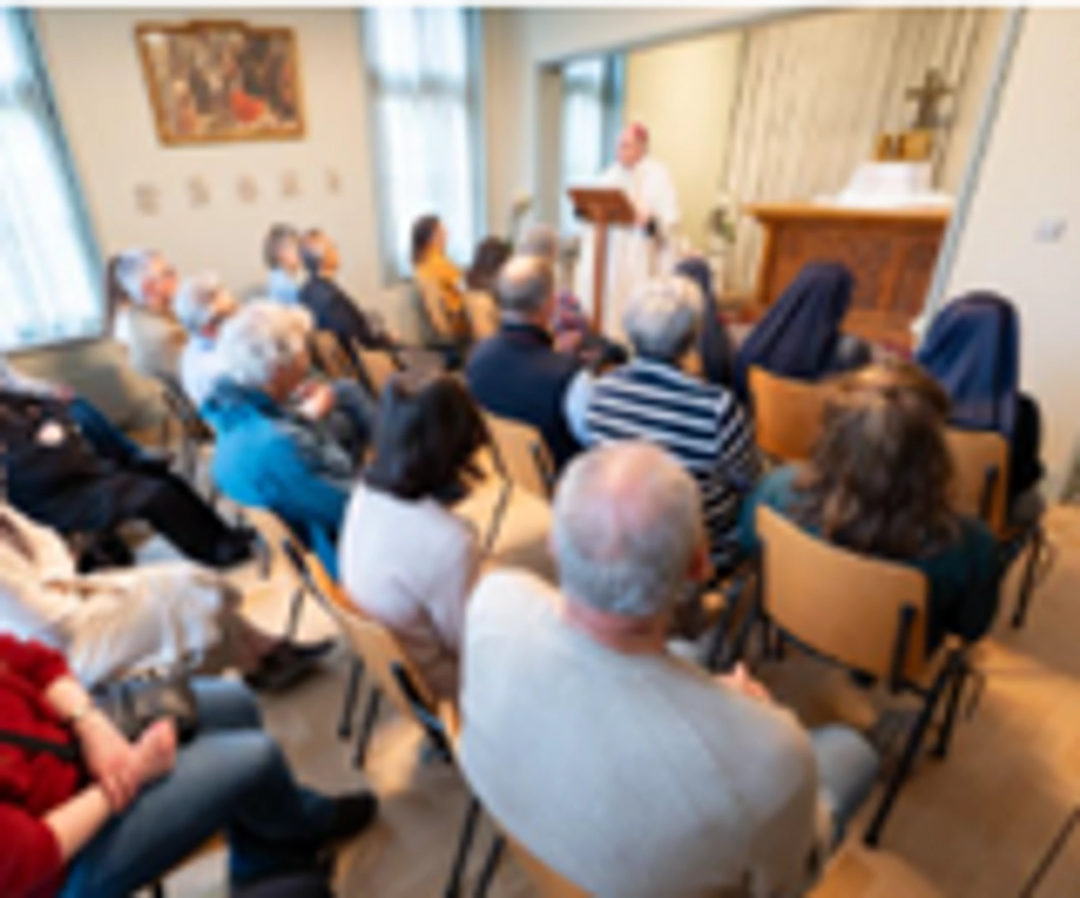A bishop in vestments speaks from a lectern to a seated audience of laypeople and nuns in a small, sunlit chapel during a religious service.