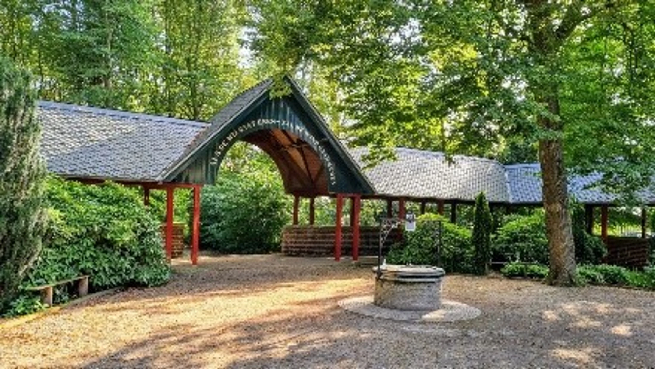 An old stone wishing well sits in a clearing in front of a long wooden covered walkway with a tiled roof, surrounded by lush green trees and bushes.