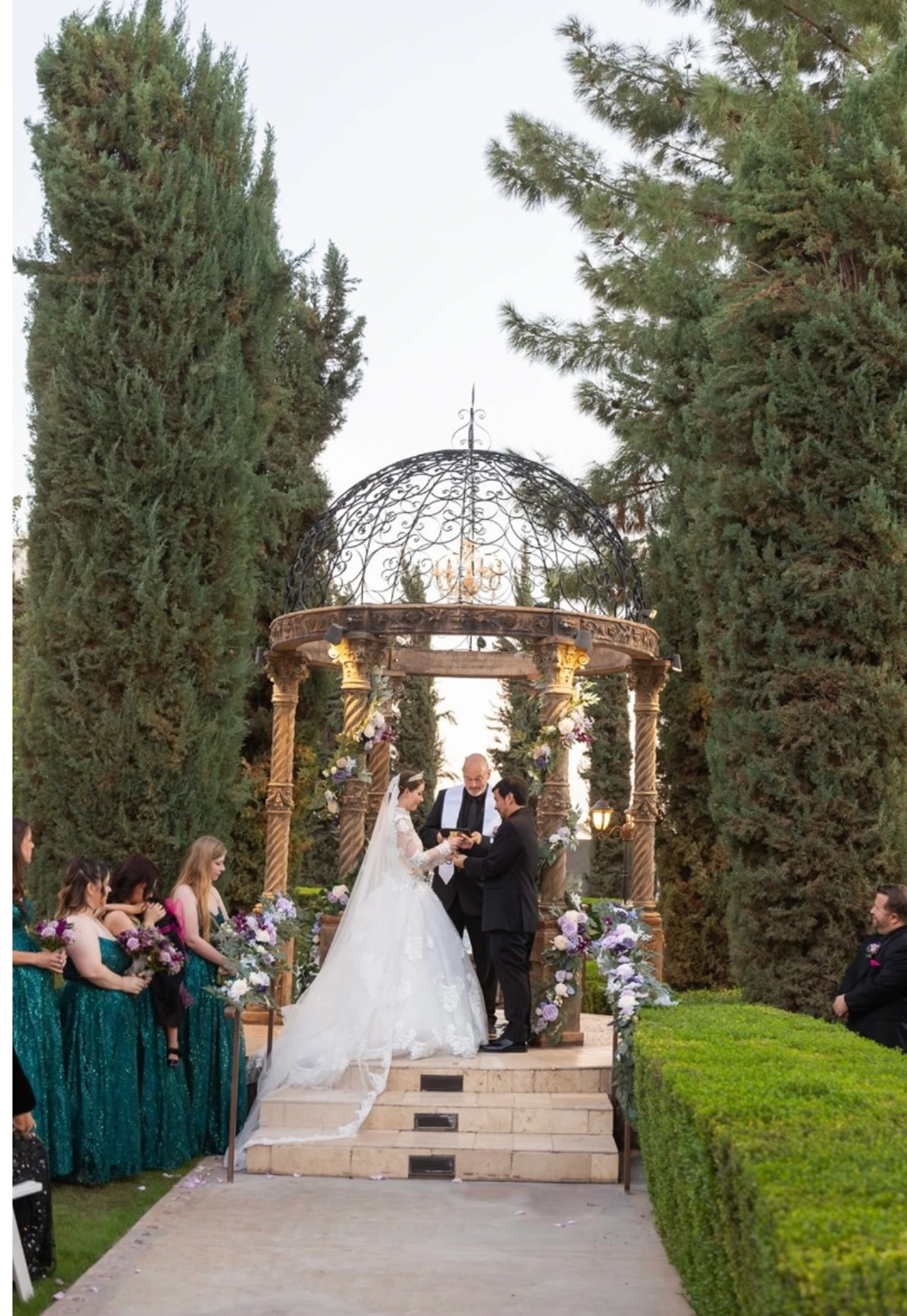 A bride and groom exchange rings under an ornate gazebo during their wedding ceremony, surrounded by tall cypress trees and their wedding party.