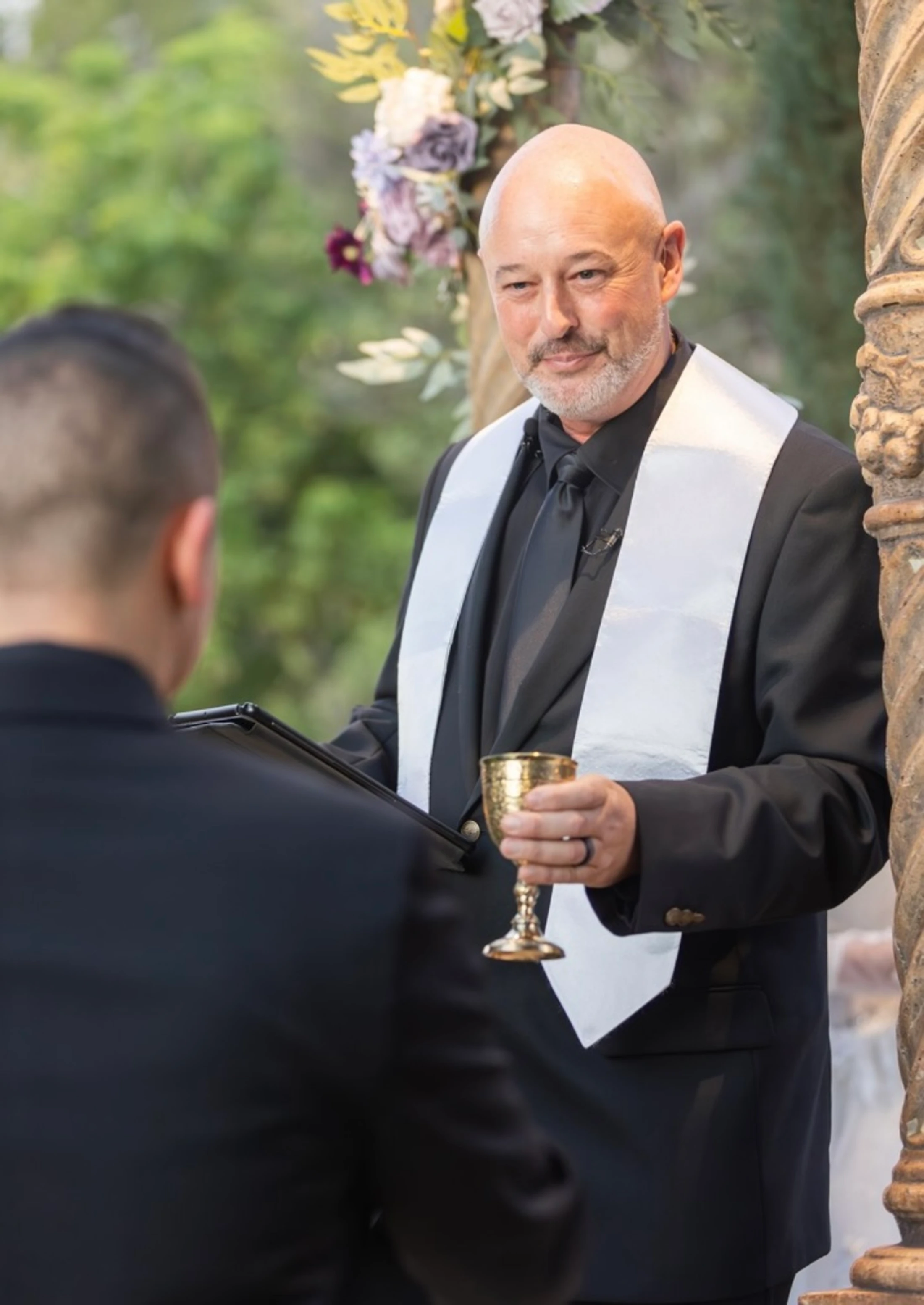 A smiling wedding officiant in a black suit and white stole holds a golden goblet while looking at the groom during an outdoor ceremony.