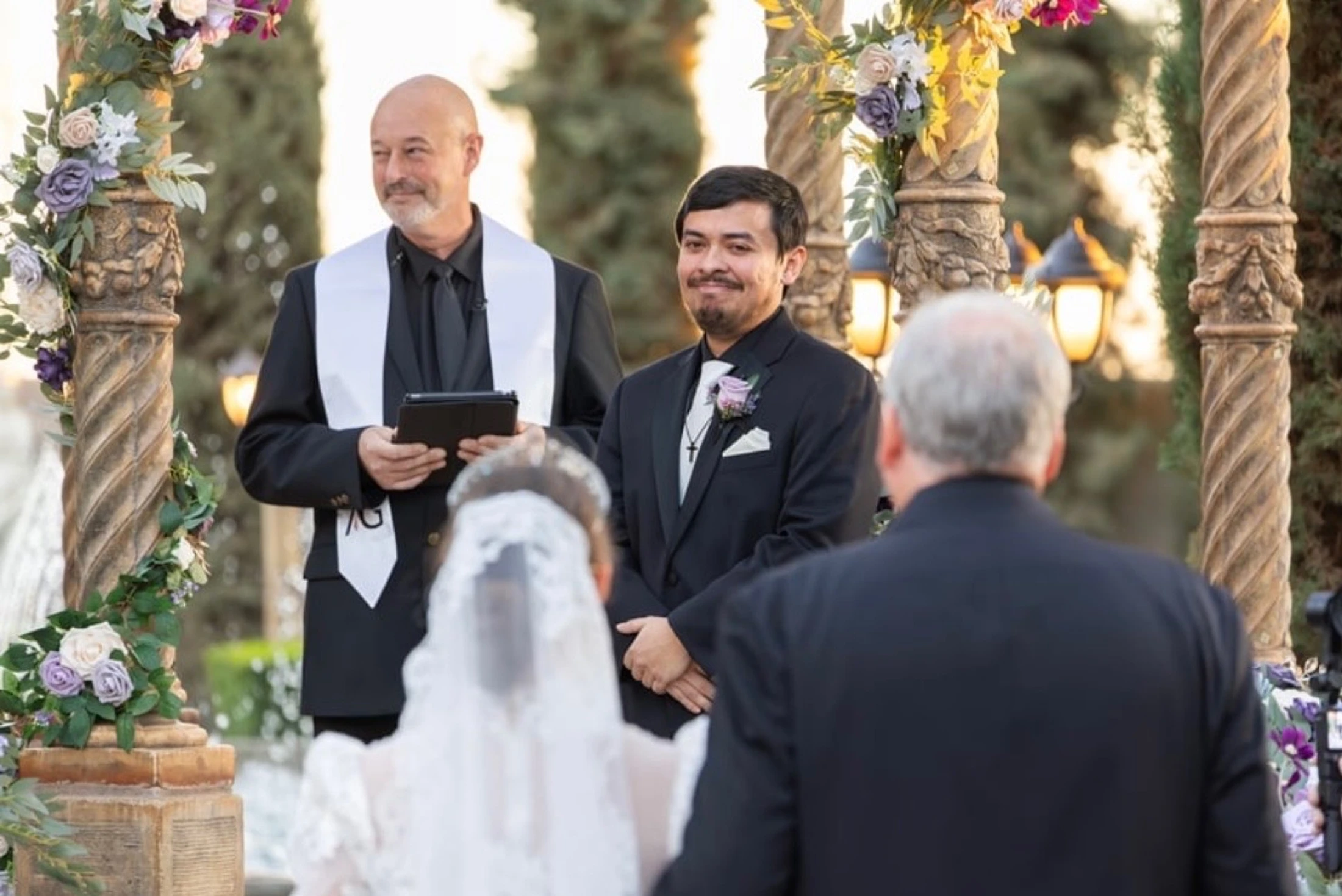 A smiling groom in a black suit watches his bride approach the altar during an outdoor wedding ceremony, with the officiant standing beside him.