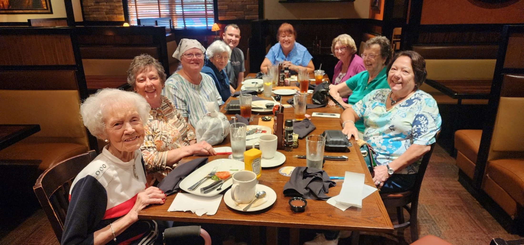 A group of nine smiling people, mostly senior women, sitting at a long wooden table in a restaurant booth, posing for a photo during their meal.