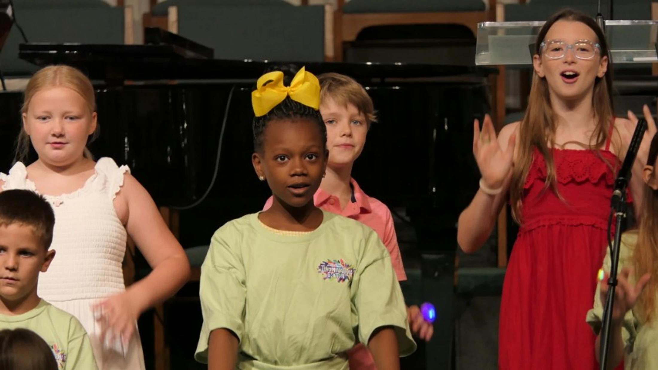 A diverse group of young children singing on a stage. A girl in a green shirt with a yellow bow is in the center, and a girl in a red dress sings beside her.