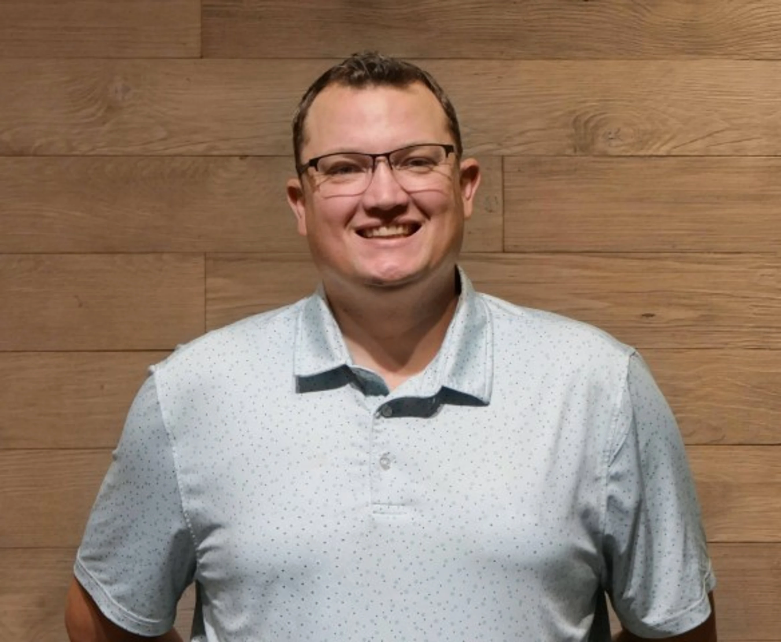 A head-and-shoulders photo of a smiling man with short brown hair and glasses, wearing a light blue polo shirt against a wood-paneled background.