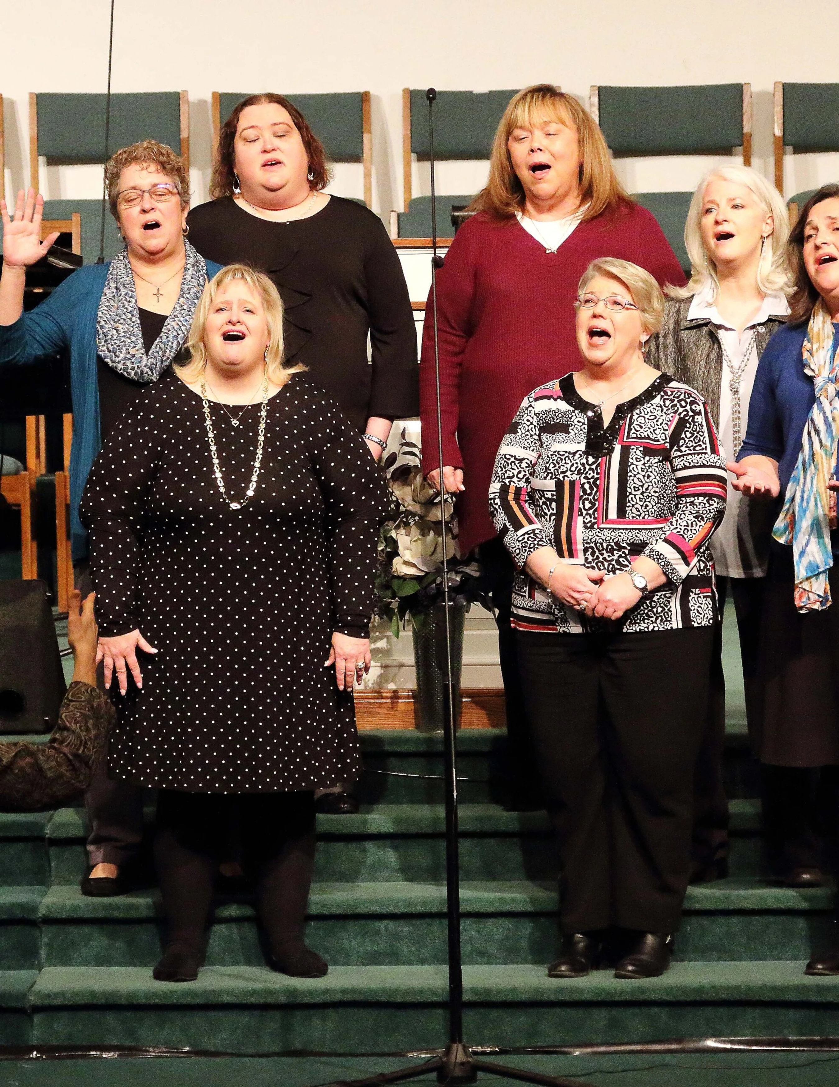 A group of seven women in a choir stand on green carpeted steps, singing with expressive faces during a performance in what appears to be a church.