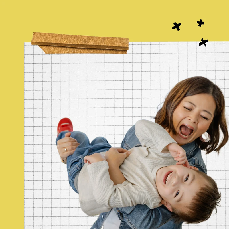 A happy mother in a denim jacket playfully holds her smiling young son sideways against a bright yellow background with The Mom Co logo.