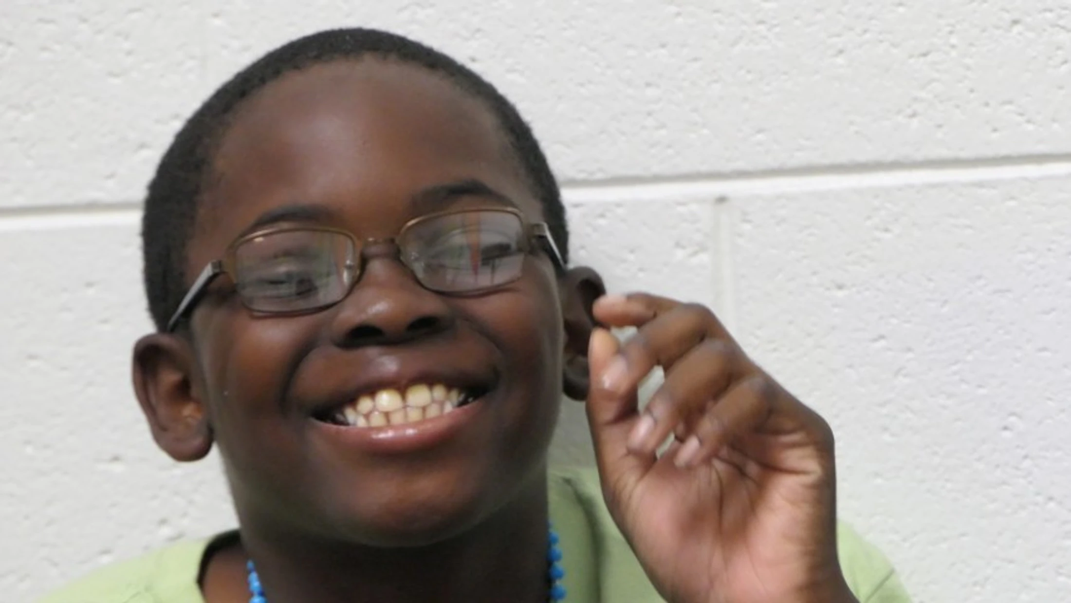 A close-up shot of a young Black boy with glasses and a green shirt, smiling broadly at the camera against a white brick wall background.