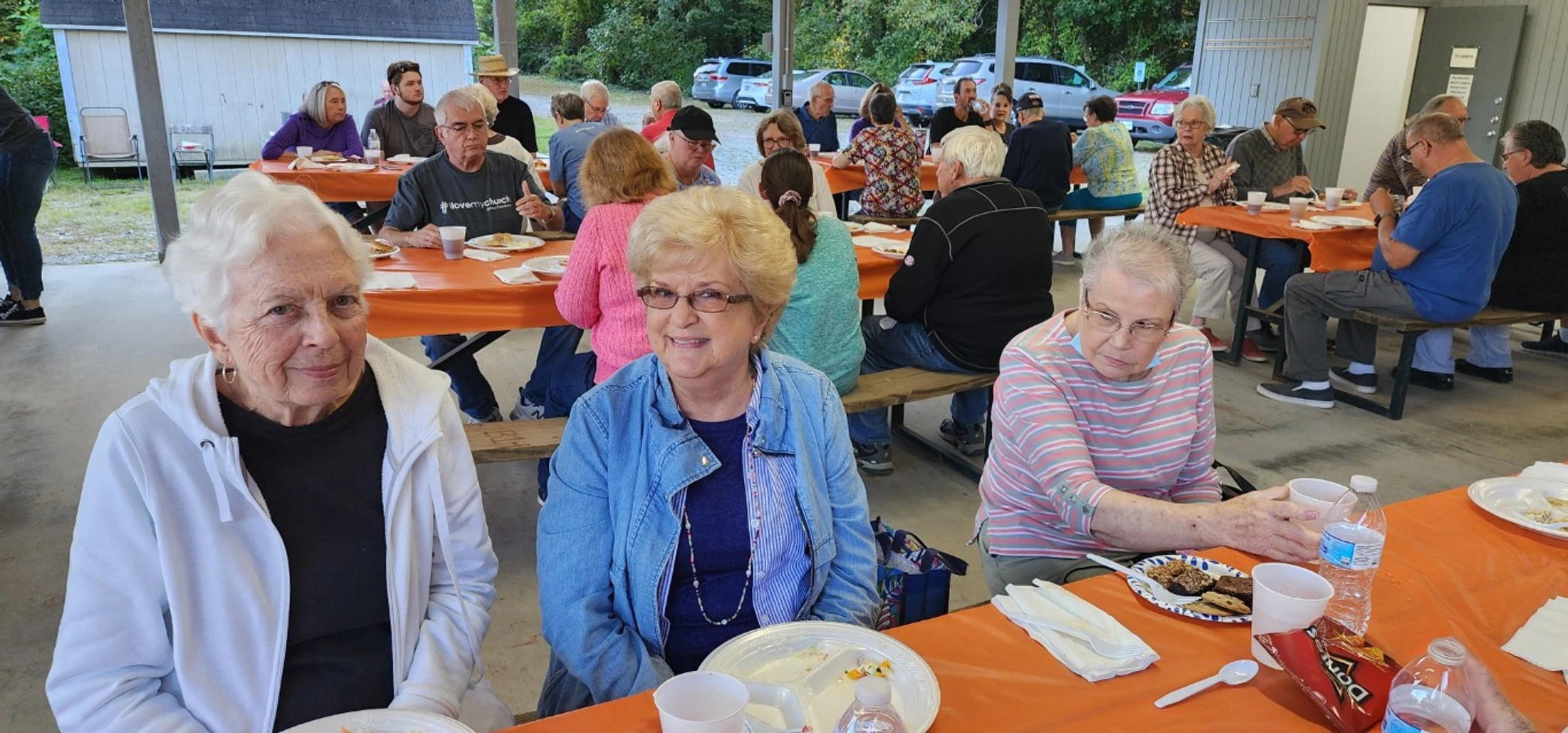 A large group of people, mostly seniors, eating and socializing at long picnic tables with orange tablecloths under a covered outdoor pavilion.