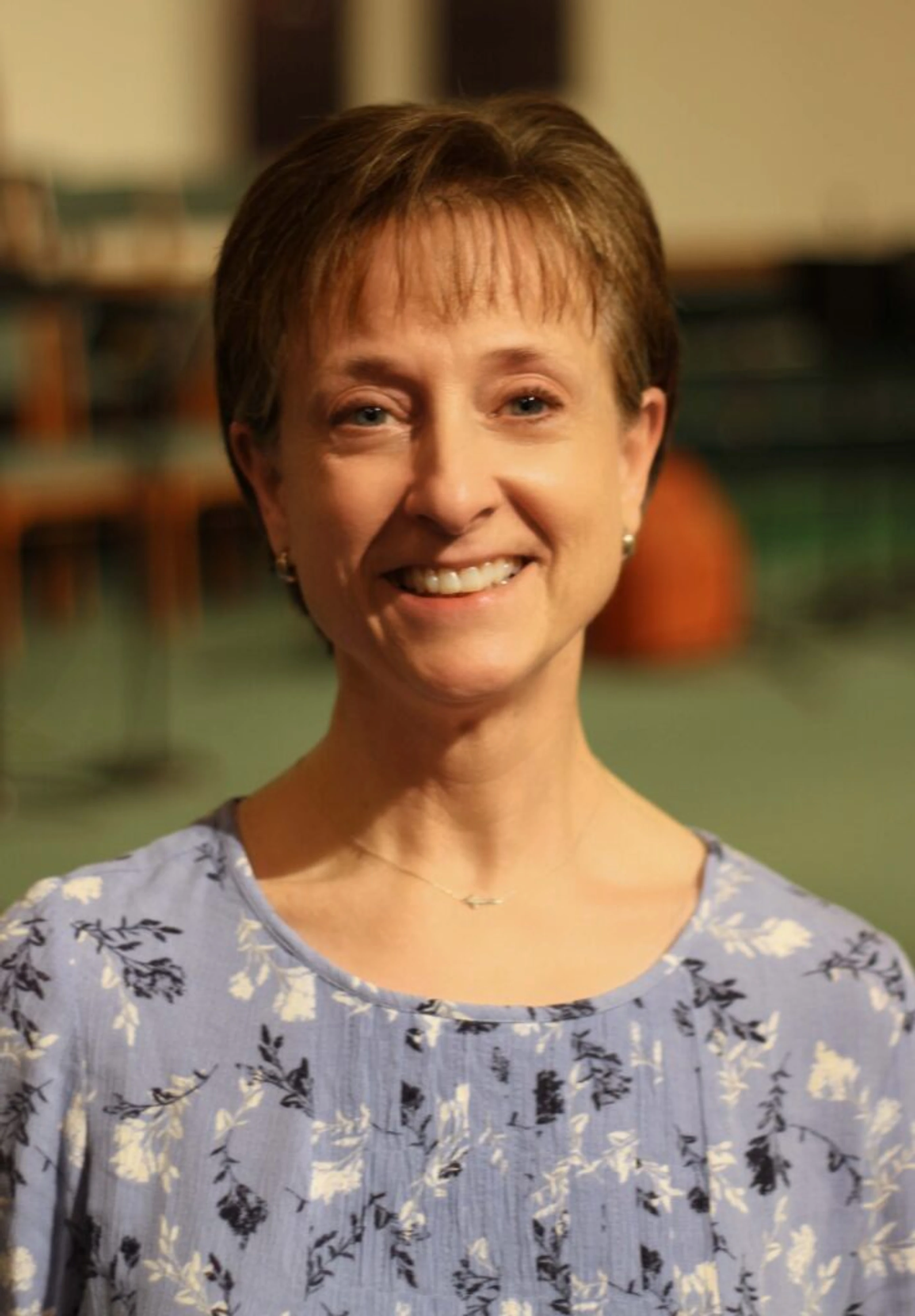 A headshot of a middle-aged woman with short brown hair and blue eyes. She is smiling at the camera and wearing a light blue floral print blouse.