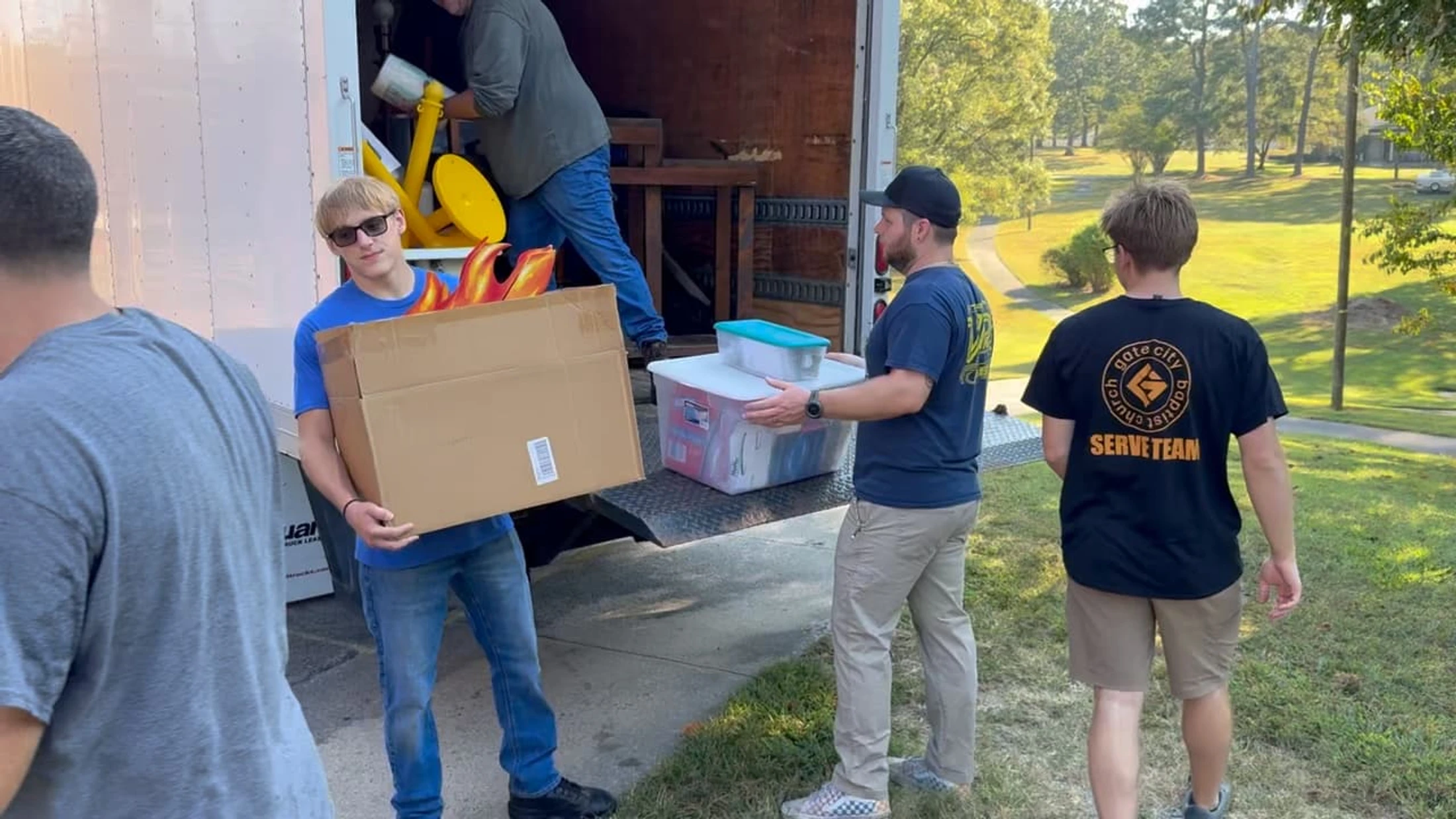 Several men from a church serve team work together to load cardboard boxes and plastic storage containers onto the back of a large moving truck.