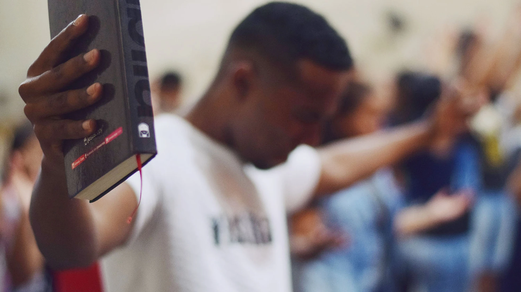 A close-up of a man's hand holding a Bible. In the soft-focus background, the man and others in a congregation are worshiping with raised hands.
