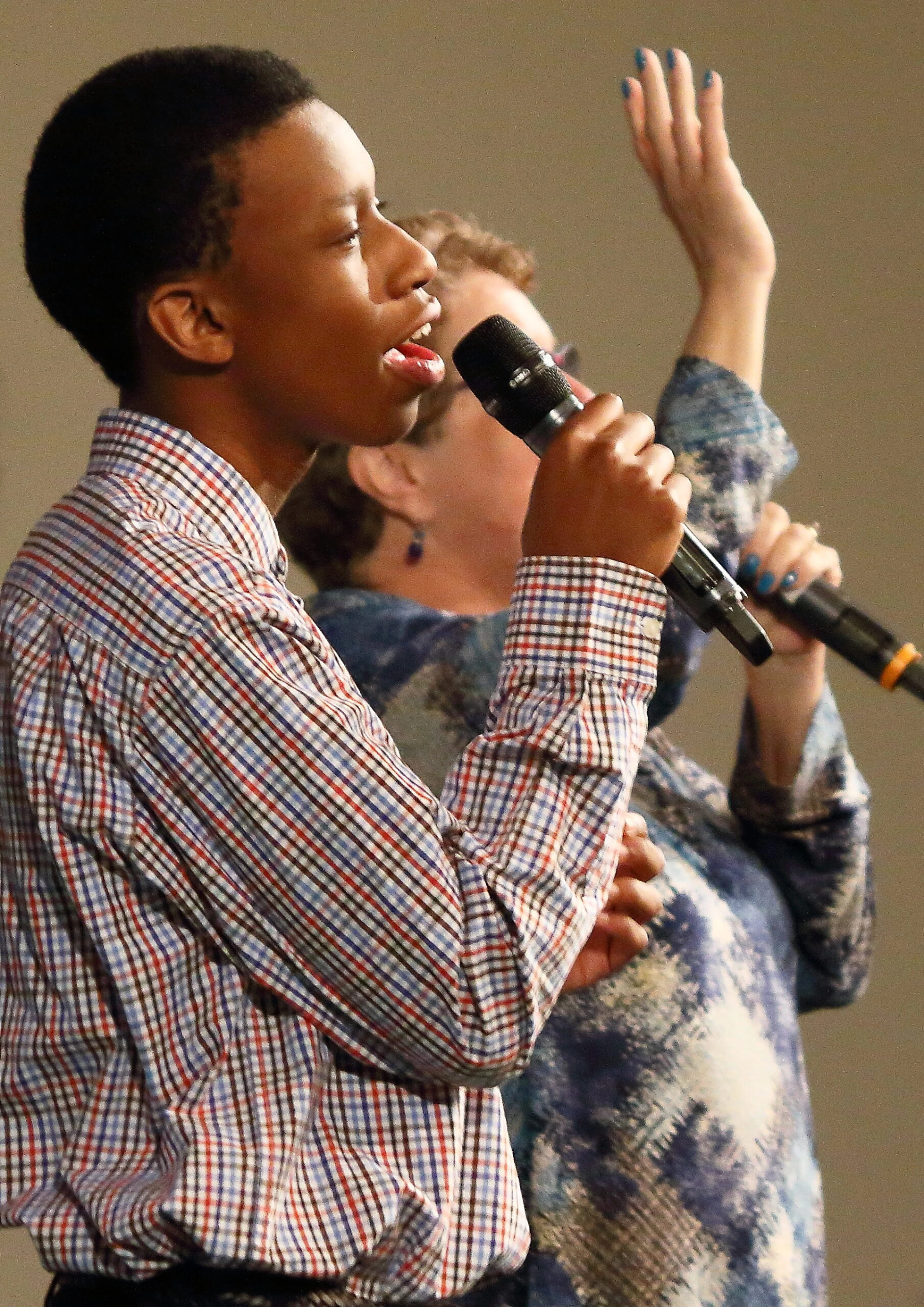 A young Black man in a plaid shirt sings into a microphone. In the background, a woman also sings with her hand raised in a gesture of worship.
