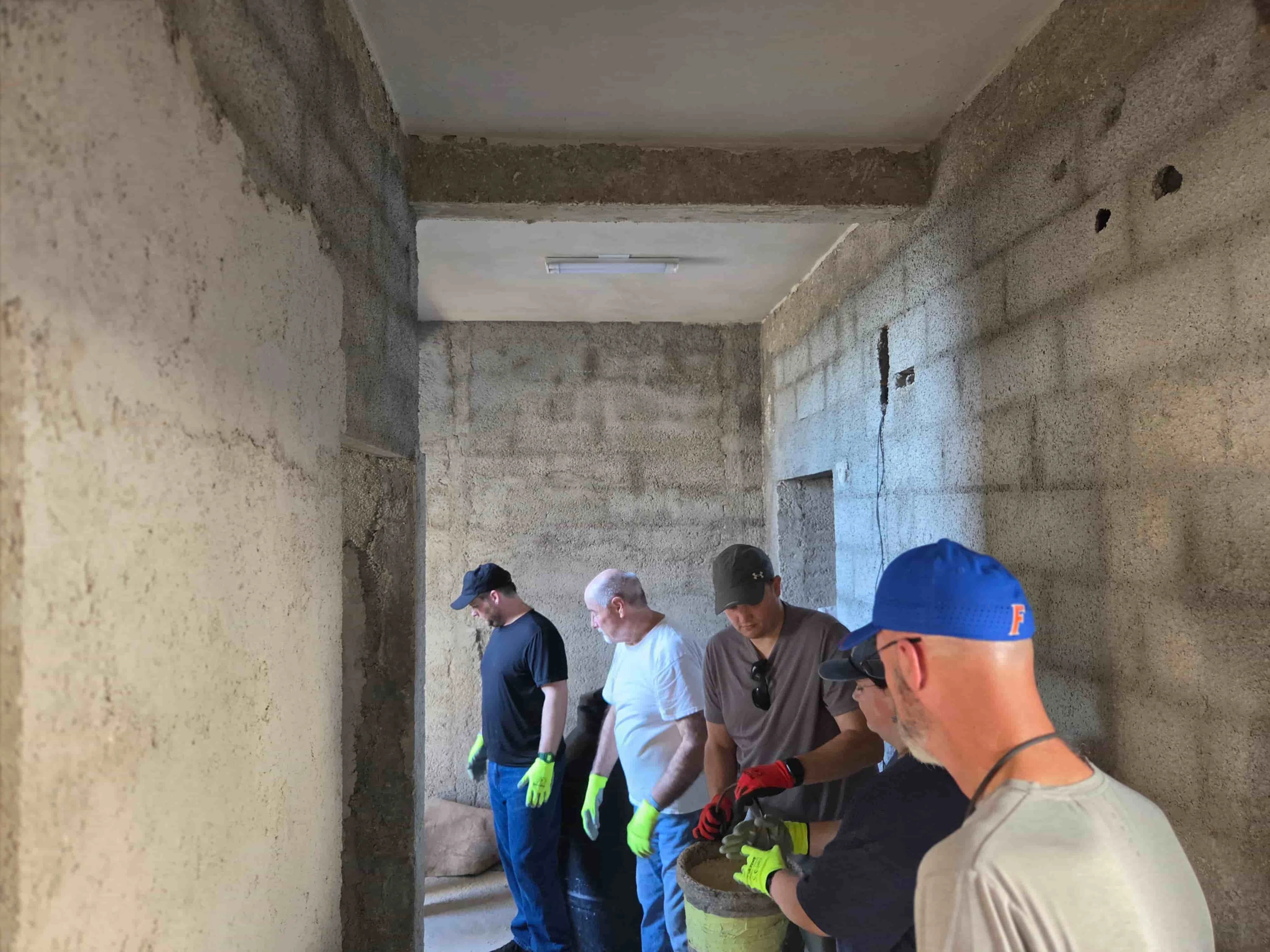 A group of five men in an unfinished concrete block hallway gather around a yellow bucket, mixing materials for a construction project.