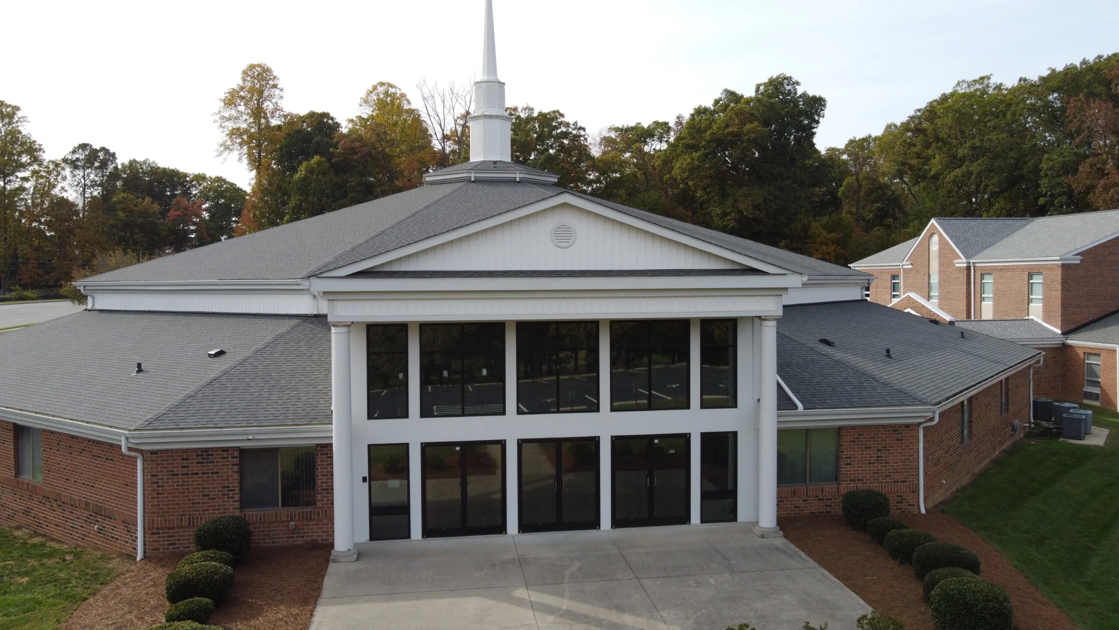 An aerial view of a modern brick church with a white facade, large dark windows, and a tall white steeple. Trees with autumn foliage are in the background.