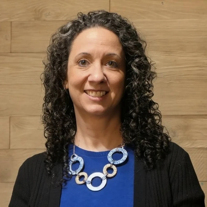 A headshot of a smiling woman with dark, curly hair against a wood-paneled wall. She wears a blue shirt, black cardigan, and a large circular necklace.