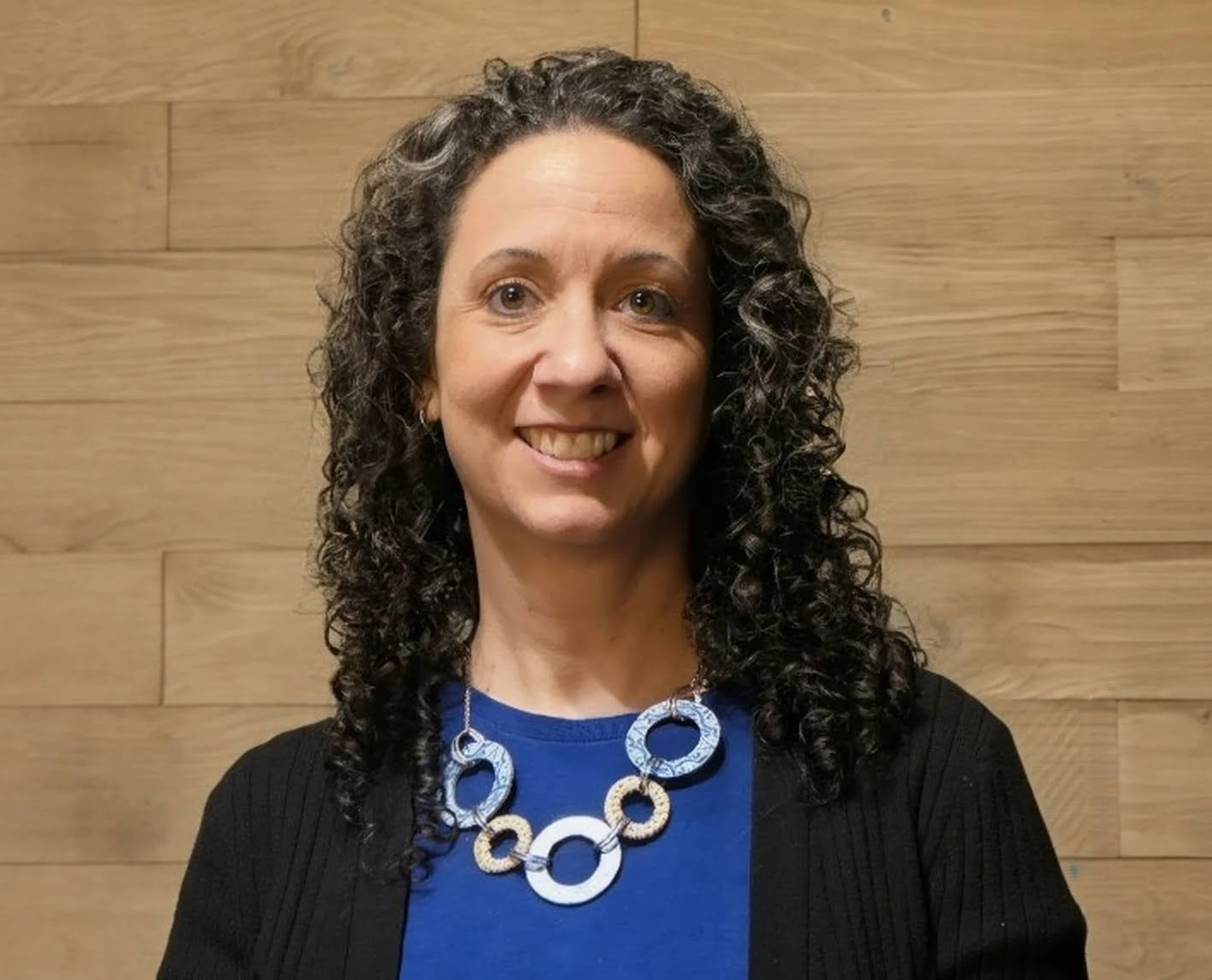 A headshot of a smiling woman with dark, curly hair against a wood-paneled wall. She wears a blue shirt, black cardigan, and a large circular necklace.