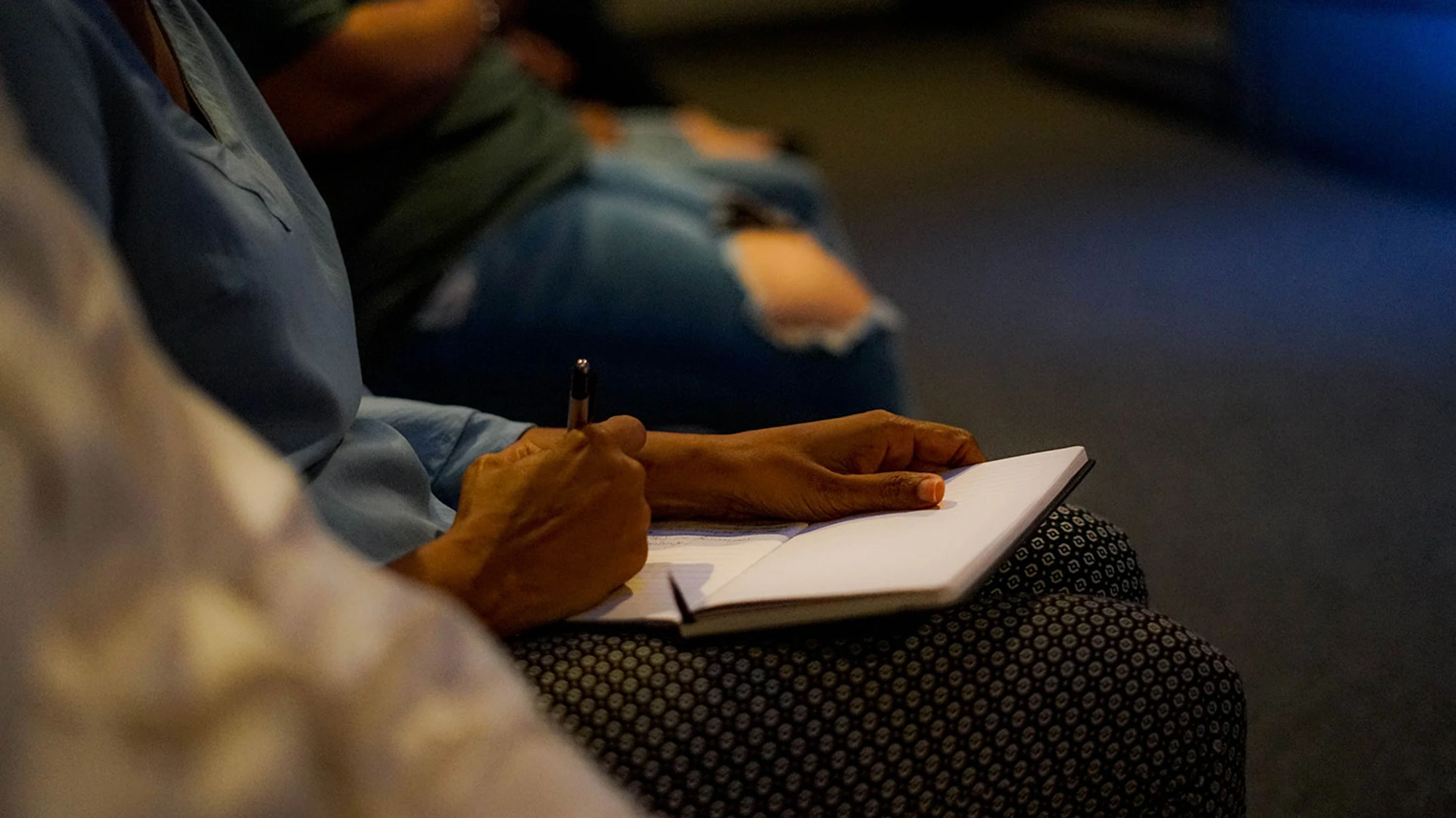A close-up of a person's hands writing with a pen in a notebook that is resting on their lap. Another person is sitting in the blurred background.