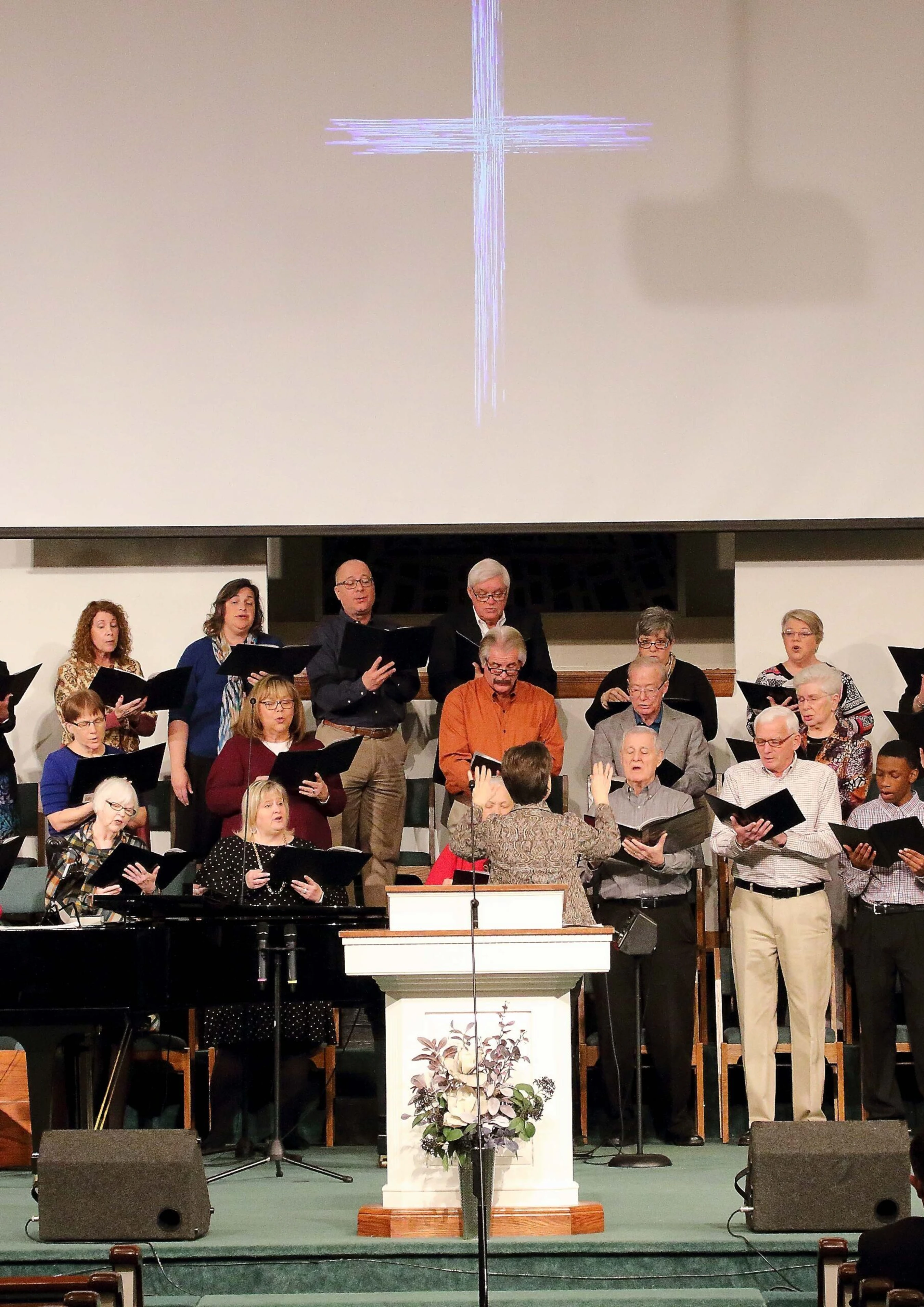 A diverse church choir of men and women of various ages sings from songbooks on a stage. A large, stylized cross is projected on the screen behind them.