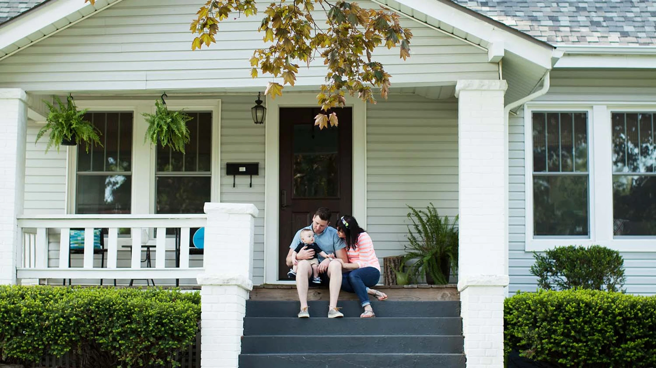 A smiling couple sits on the dark gray steps of their light-colored house, holding their baby boy on their laps under a leafy tree branch.