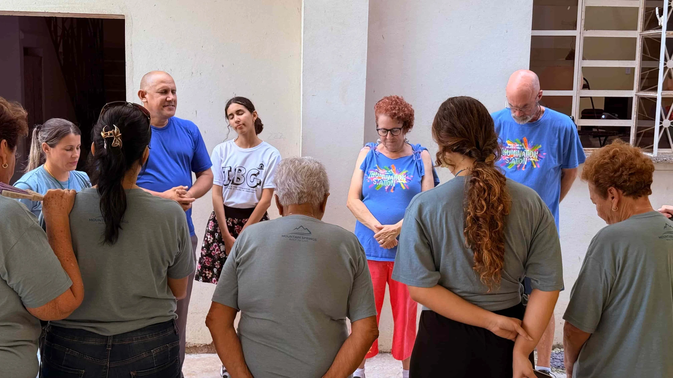 A diverse group of men and women stand in a circle with their heads bowed and eyes closed in a moment of prayer or quiet reflection outdoors.