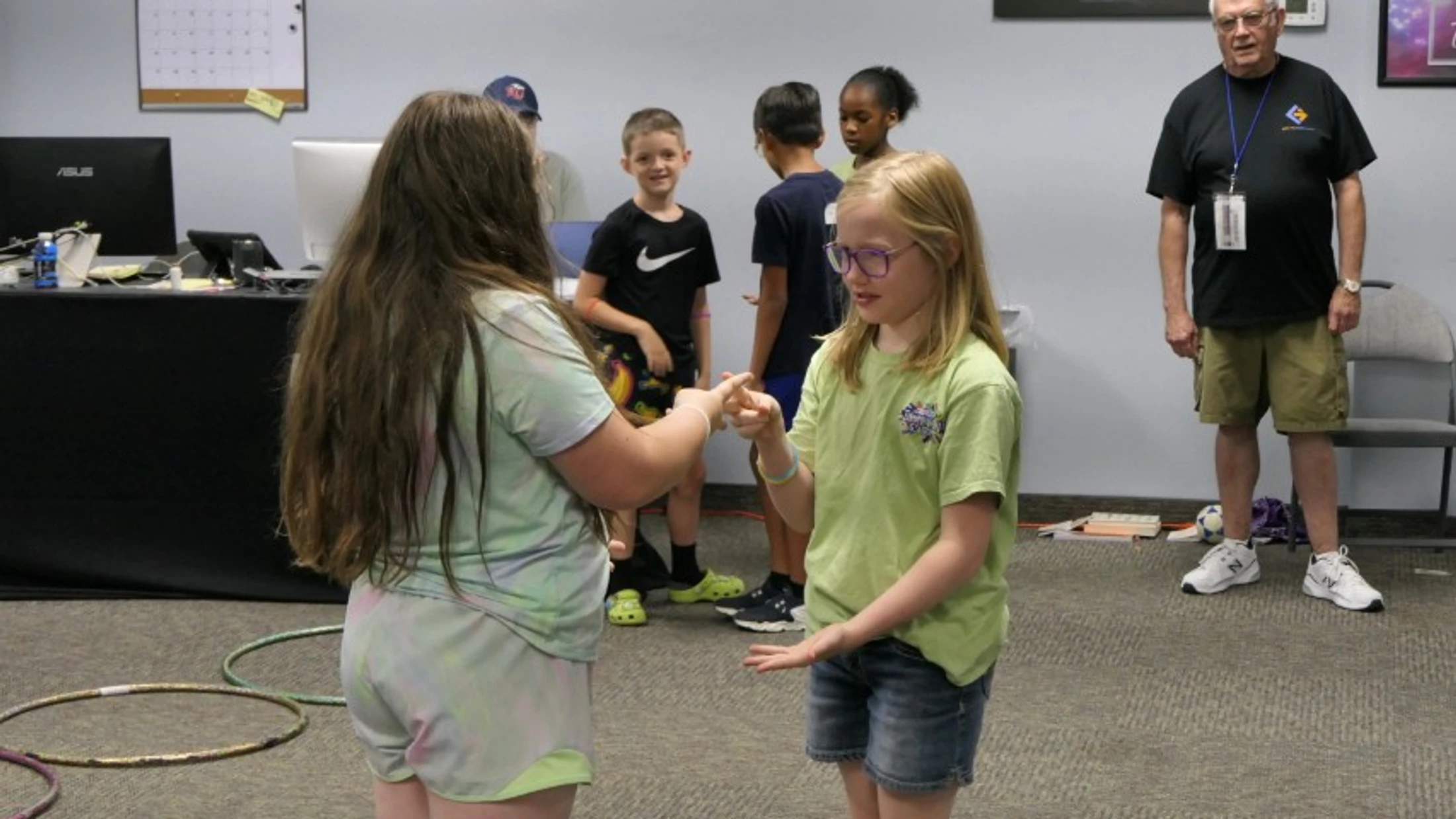 Two young girls play a hand game while other children and an adult watch in a classroom. Hula hoops are on the carpeted floor in the foreground.