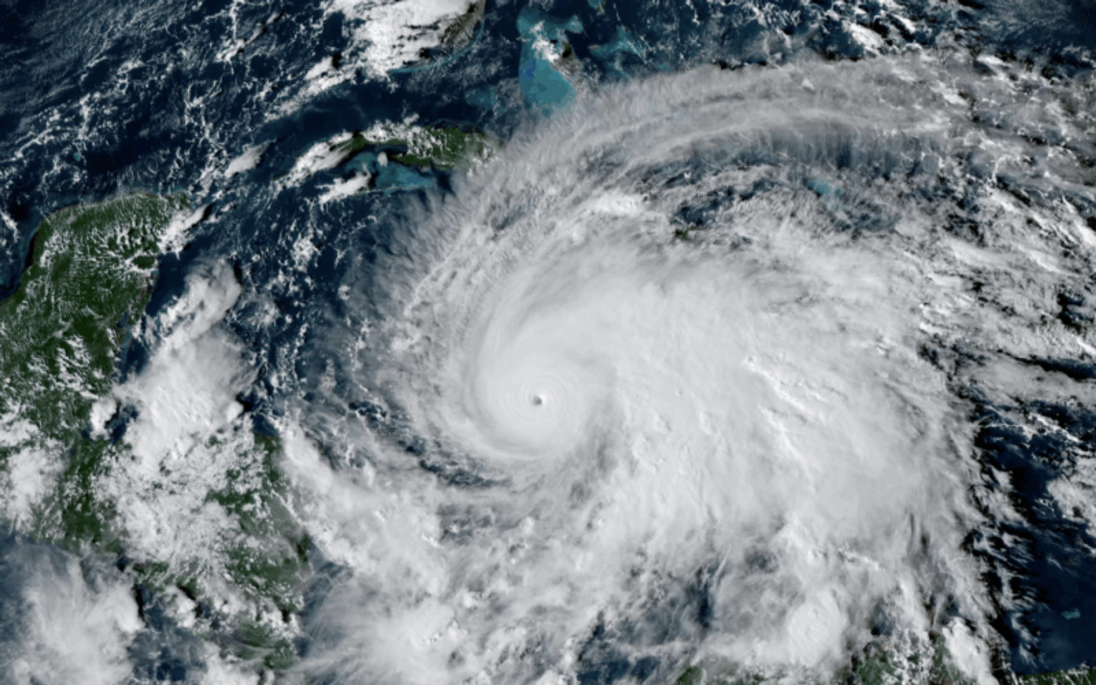 A satellite view of a massive, well-formed hurricane with a clear eye at its center, churning over the blue ocean and near green landmasses.