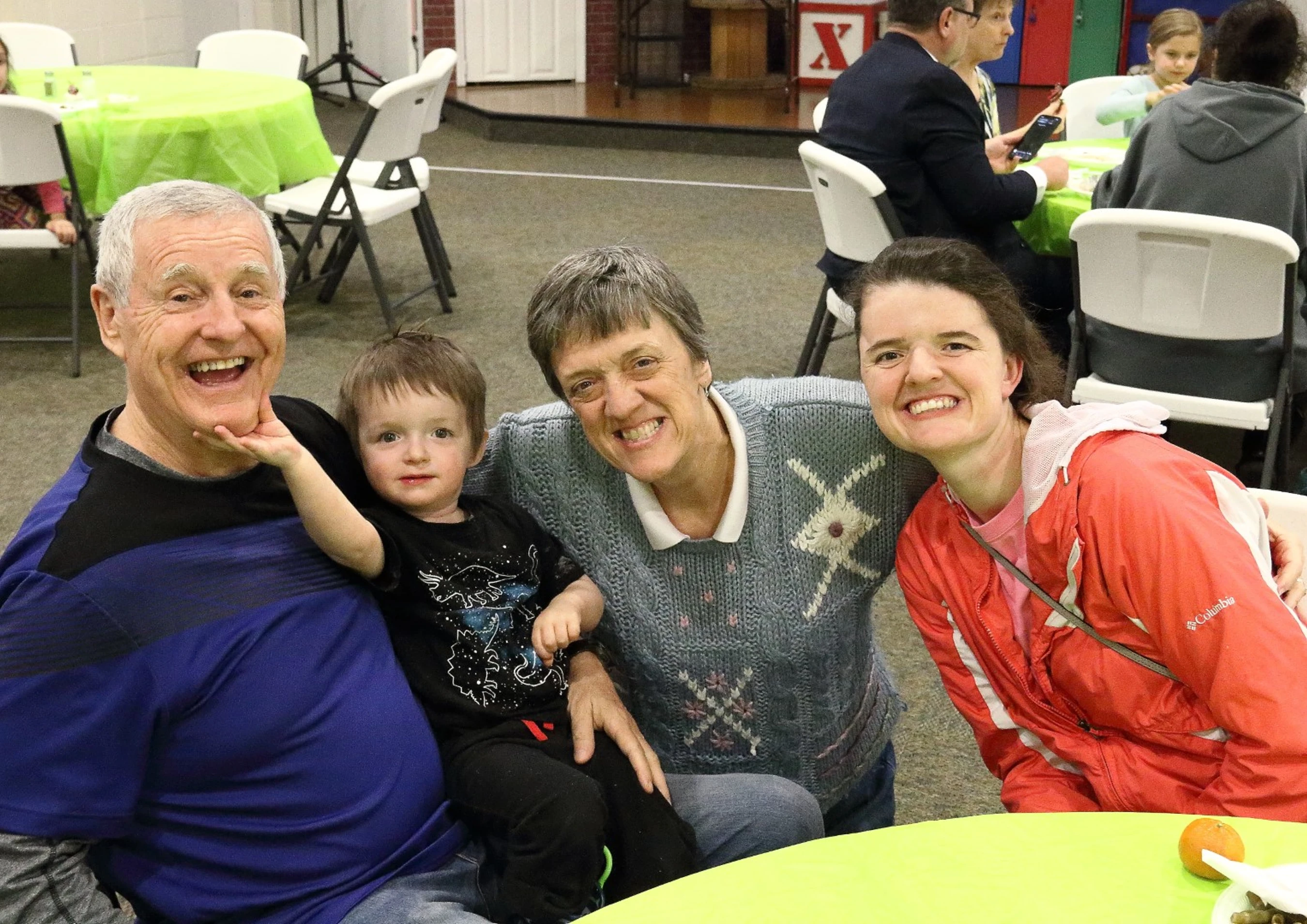 A happy family of four poses for a photo. A grandfather holds his grandson, who touches his chin, next to a grandmother and a younger woman, all smiling.