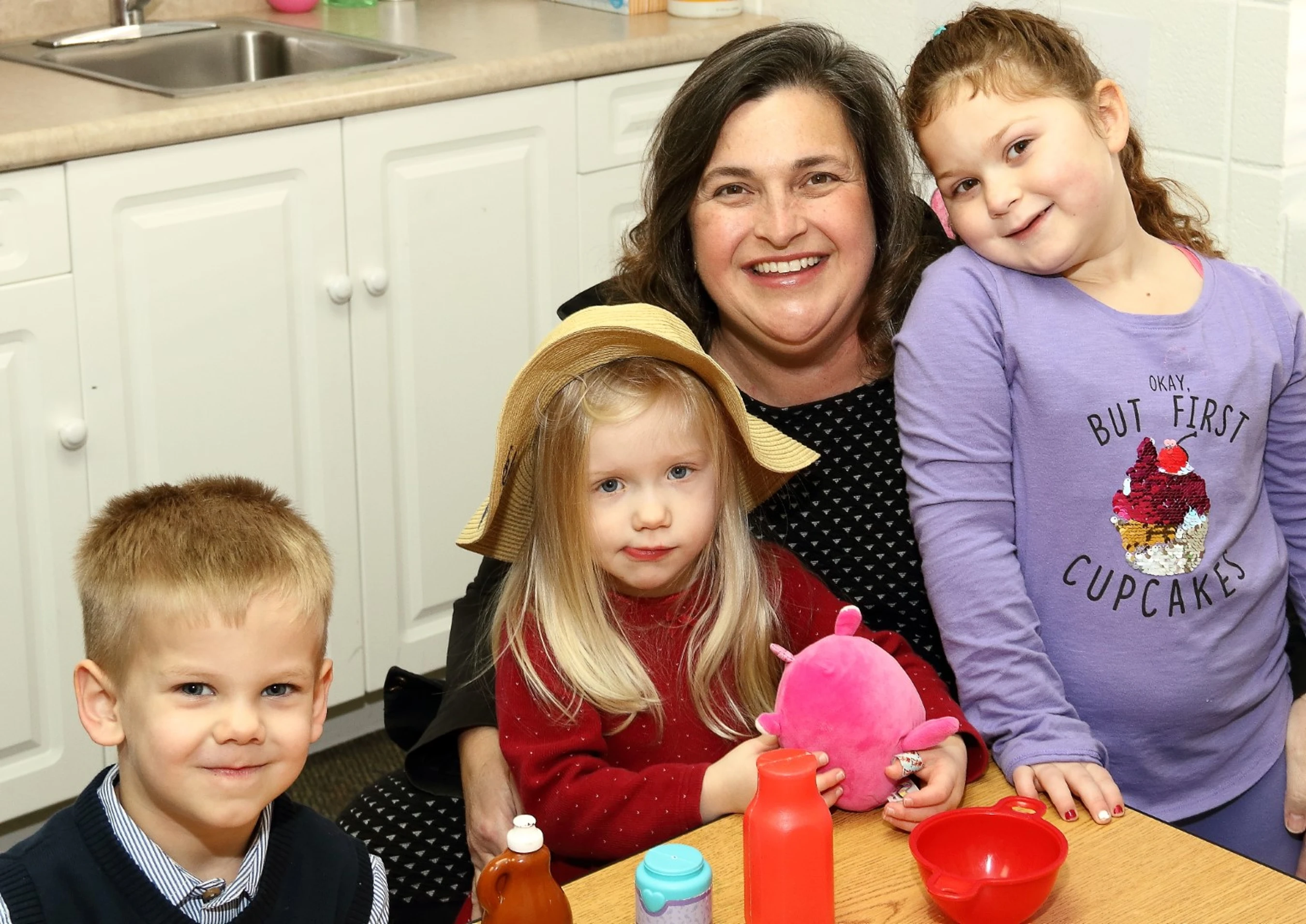 A smiling woman sits with three young children at a table. A boy is on the left, a girl in a hat is in the center, and another girl leans in from the right.